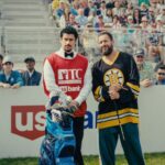 Adam Sandler as Happy Gilmore in his Boston Bruins hockey jersey, holding a golf club, standing next to his caddy on a golf course with spectators in the background.