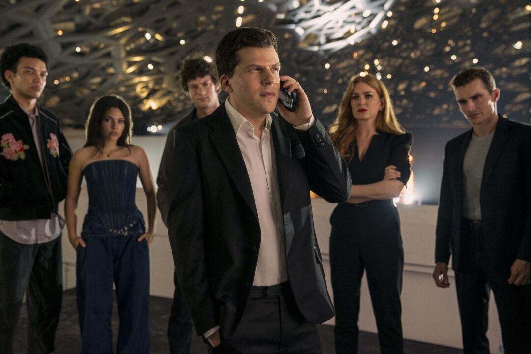 J. Daniel Atlas (Jesse Eisenberg) on a phone, surrounded by several other cast members, including Isla Fisher as Henley, and others, in what appears to be a large, modern, possibly event space, in Now You See Me, Now You Don't.