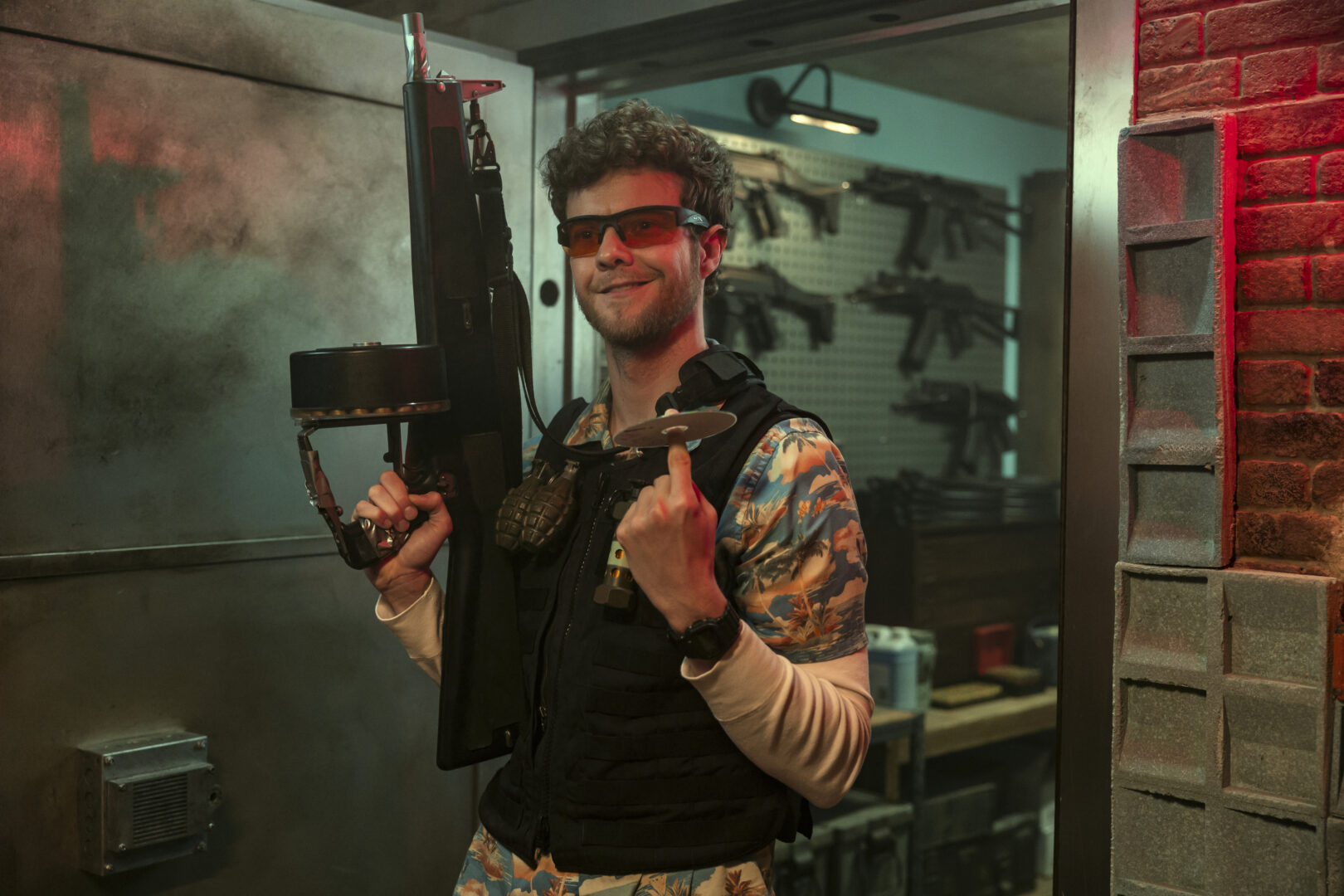 Jack Quaid wearing sunglasses and a Hawaiian shirt, holding a large firearm with a drum magazine, in a room full of weapons from Heads of State.
