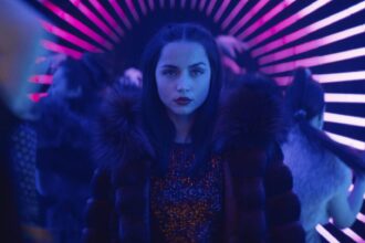 Ana de Armas as the character Rooney in Ballerina, wearing a fur-collared coat and a sequined top, with a serious expression, standing in a vibrant, neon-lit tunnel with pink and blue stripes.