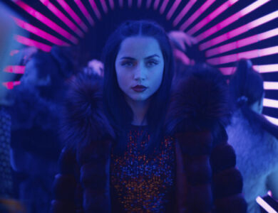 Ana de Armas as the character Rooney in Ballerina, wearing a fur-collared coat and a sequined top, with a serious expression, standing in a vibrant, neon-lit tunnel with pink and blue stripes.