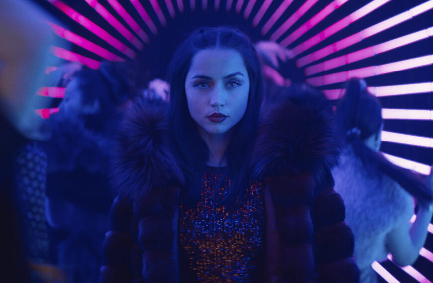 Ana de Armas as the character Rooney in Ballerina, wearing a fur-collared coat and a sequined top, with a serious expression, standing in a vibrant, neon-lit tunnel with pink and blue stripes.