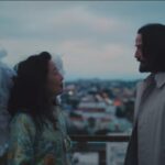 Keanu Reeves, with large white wings attached to his back, looking intently at a woman with dark hair, on a rooftop or balcony overlooking a cityscape at dusk, from the movie Good Fortune.