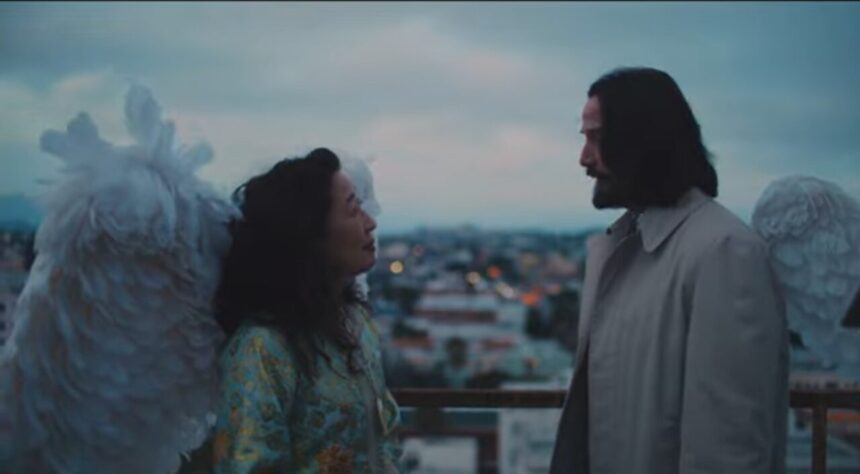 Keanu Reeves, with large white wings attached to his back, looking intently at a woman with dark hair, on a rooftop or balcony overlooking a cityscape at dusk, from the movie Good Fortune.