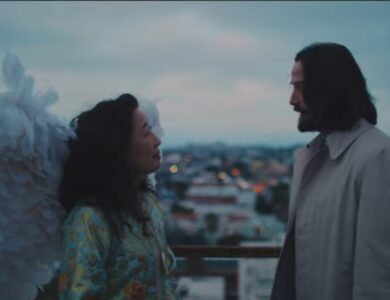 Keanu Reeves, with large white wings attached to his back, looking intently at a woman with dark hair, on a rooftop or balcony overlooking a cityscape at dusk, from the movie Good Fortune.