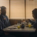 Three individuals, two men in what appear to be work uniforms and a woman eating, seated in a diner booth with venetian blinds on the windows, from the movie The Pickup.