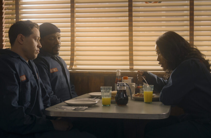 Three individuals, two men in what appear to be work uniforms and a woman eating, seated in a diner booth with venetian blinds on the windows, from the movie The Pickup.
