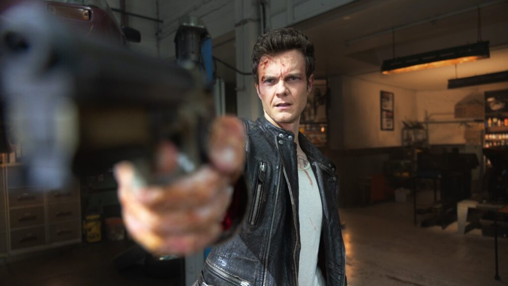 Jack Quaid as his character in Novocaine, wearing a leather jacket and white t-shirt, with blood smears on his face, holding a revolver and looking determined in a dimly lit garage or industrial setting.