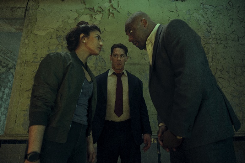 From Heads of State, Idris Elba, John Cena, and Priyanka Chopra Jonas stand in a tense, dilapidated room, looking at each other with serious expressions, Cena positioned slightly behind the other two.