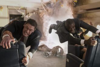 John Cena and Idris Elba, dressed in suits, struggle to hold onto seats in a rapidly disintegrating airplane cabin filled with smoke and debris, their faces strained.
