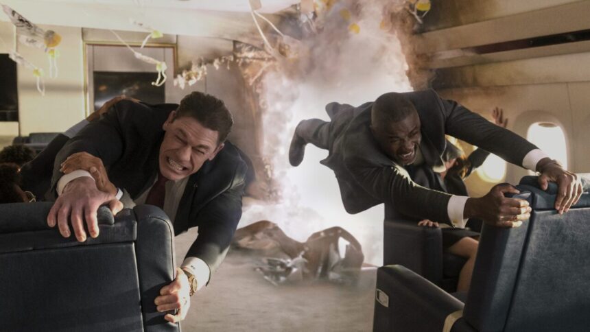 John Cena and Idris Elba, dressed in suits, struggle to hold onto seats in a rapidly disintegrating airplane cabin filled with smoke and debris, their faces strained.