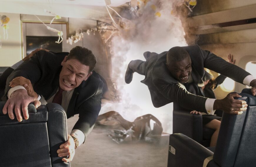 John Cena and Idris Elba, dressed in suits, struggle to hold onto seats in a rapidly disintegrating airplane cabin filled with smoke and debris, their faces strained.