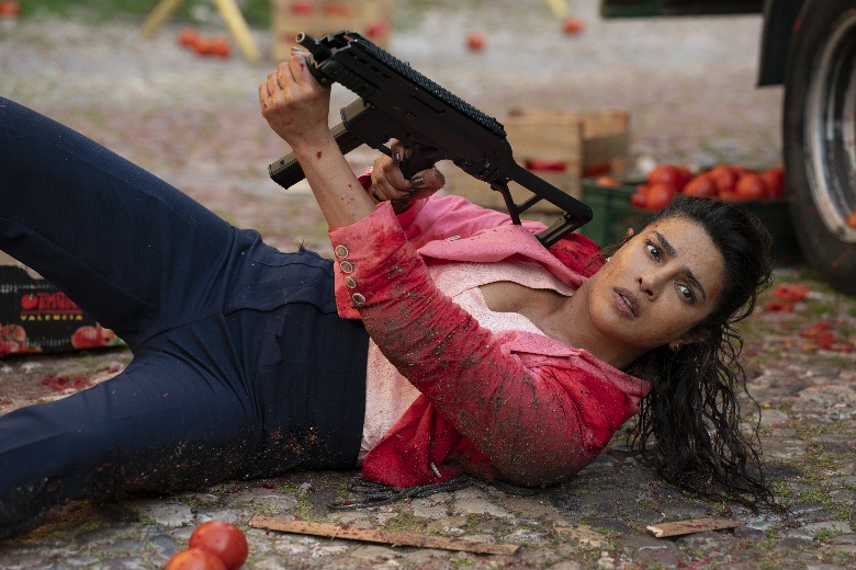 Priyanka Chopra Jonas in a pink jacket, lying on her side amidst scattered tomatoes, aiming a black automatic weapon with a determined expression.