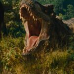 A massive T-Rex roars, jaws wide, at a person with their back to the camera, amidst lush green foliage in a scene from Jurassic World Rebirth.
