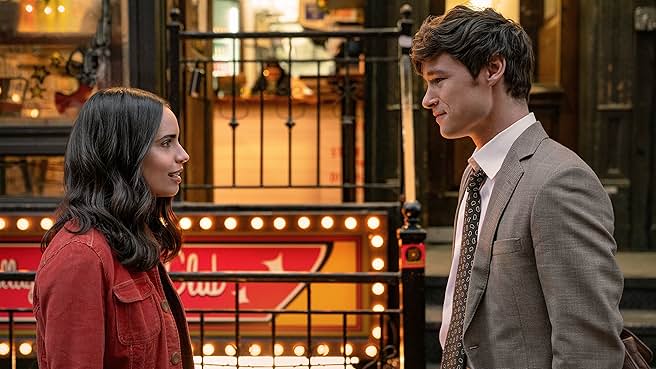 Sofia Carson and Kyle Allen as characters in 'The Life List' standing on a city street, looking at each other with soft smiles, surrounded by storefront lights.