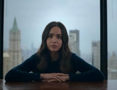 Sofia Carson as the protagonist of 'The Life List' sitting at a large table in front of a window overlooking a city skyline, looking thoughtful.