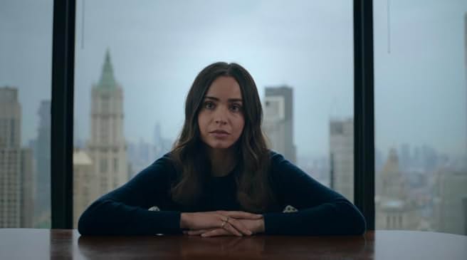 Sofia Carson as the protagonist of 'The Life List' sitting at a large table in front of a window overlooking a city skyline, looking thoughtful.