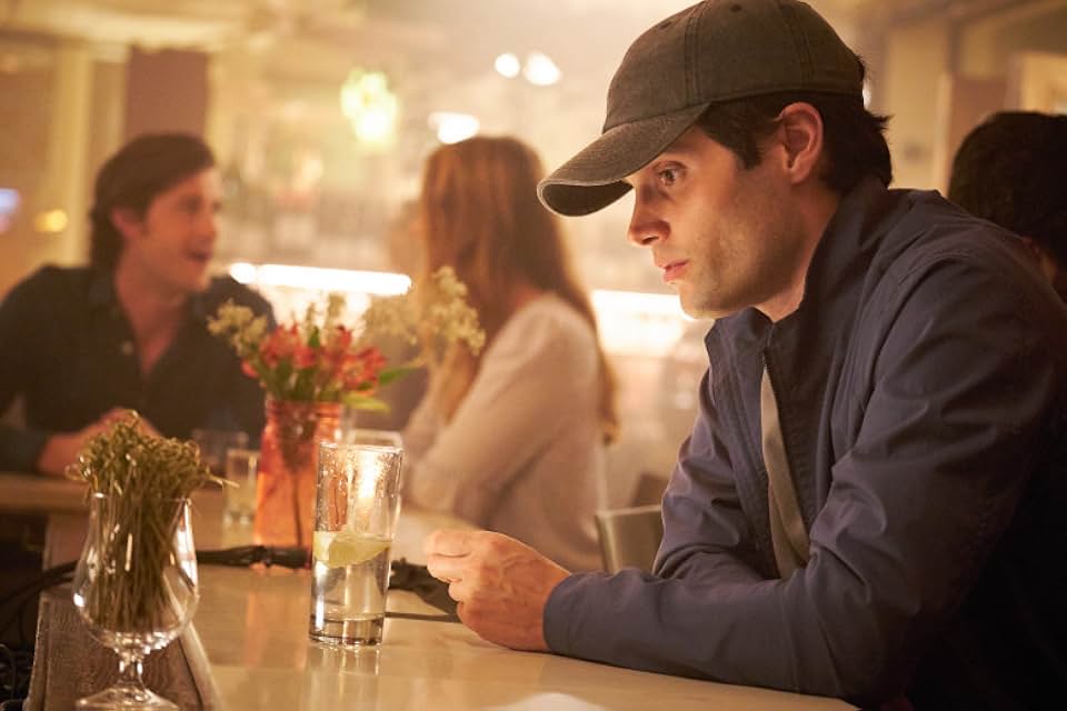 Penn Badgley as Joe Goldberg wearing a baseball cap, observing someone intently from a bar stool in a dimly lit setting.