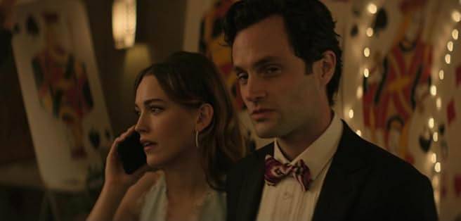 Joe Goldberg (Penn Badgley) and Love Quinn (Victoria Pedretti) dressed up, possibly at a party, with playing card-themed decor in the background, from Season 3 of Netflix's YOU.