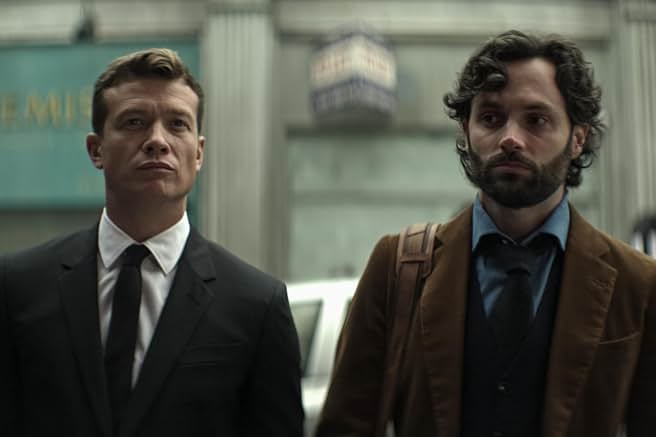 Penn Badgley as Joe Goldberg (on the right) with a brown jacket and bag, standing next to a man in a suit, on a city street in London from Season 4 of Netflix's YOU.