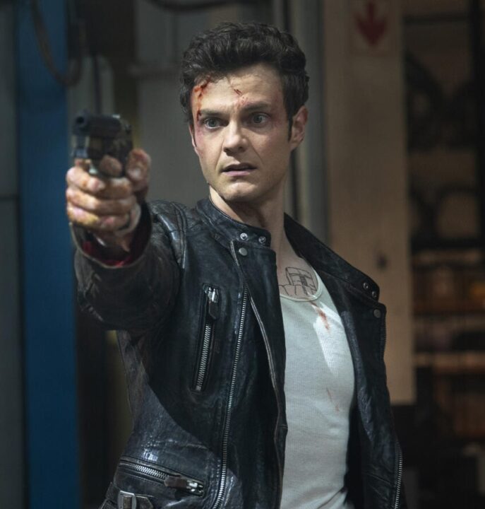 Jack Quaid as his character in Novocaine, wearing a leather jacket and white t-shirt, with blood smears on his face, holding a revolver and looking determined in a dimly lit garage or industrial setting.