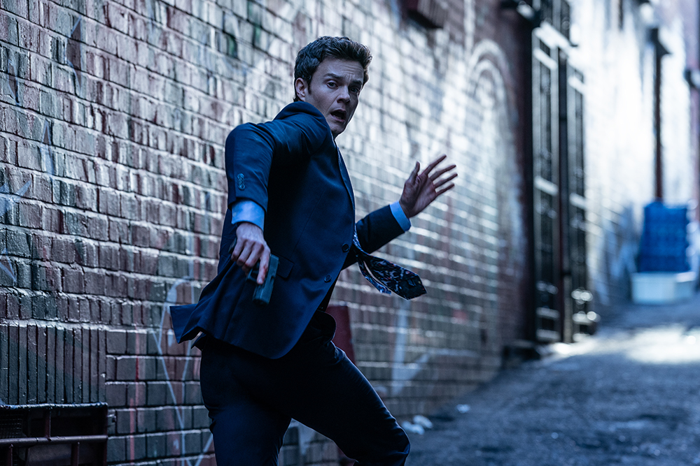Jack Quaid as his character in Novocaine, wearing a dark suit and tie, running frantically down a narrow brick alleyway while looking back over his shoulder with a surprised expression.
