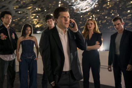 J. Daniel Atlas (Jesse Eisenberg) on a phone, surrounded by several other cast members, including Isla Fisher as Henley, and others, in what appears to be a large, modern, possibly event space, in Now You See Me, Now You Don't.