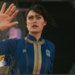 Lucy (Ella Purnell) from Fallout Season 1, wearing a Vault Suit, waving with a look of awe or slight trepidation as she exits a Vault into the wasteland.