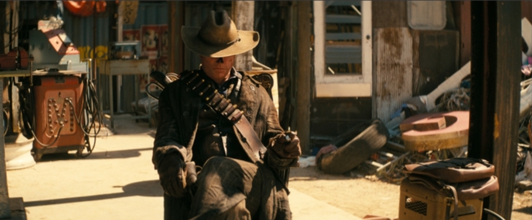 The Ghoul (Walton Goggins) from Fallout Season 1, seated in a chair in the wasteland, wearing a cowboy hat and duster, looking stern.
