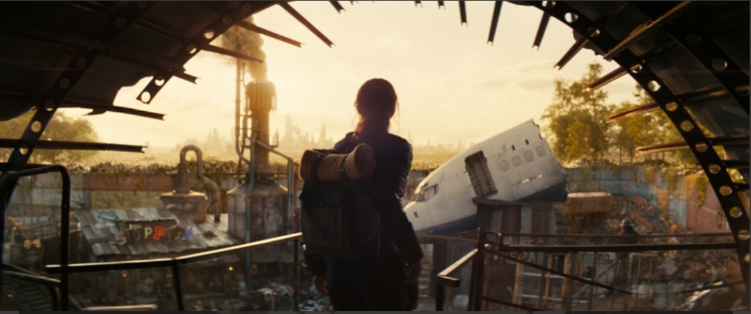 Lucy (Ella Purnell) from Fallout Season 1, seen from behind, looking out at the vast, desolate wasteland with a crashed airplane in the distance, showing the scale of destruction.