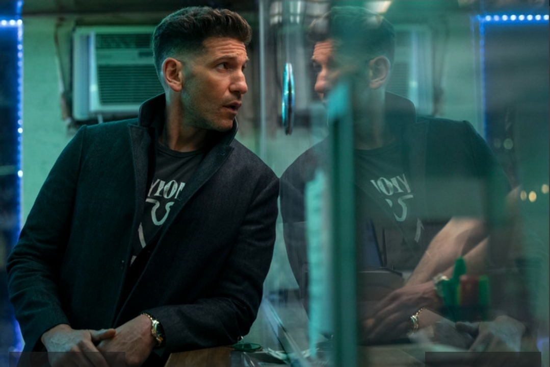 Jon Bernthal as Braxton, looking to the side, with his reflection visible in a glass surface in a hotel lobby, bathed in blue and purple neon lights.