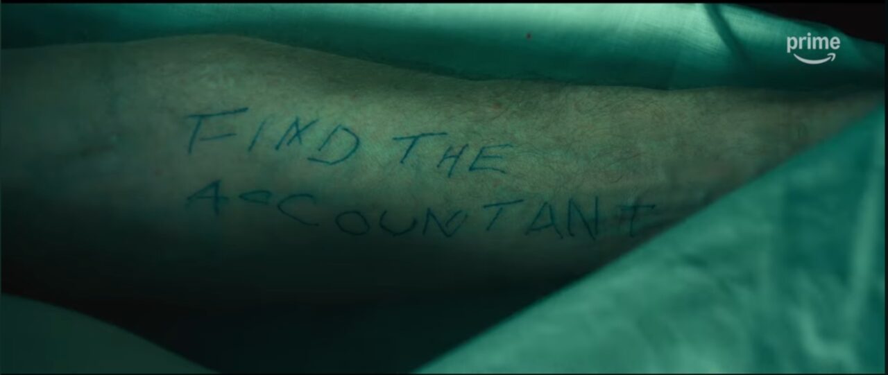 A close-up of a forearm with "FIND THE ACCOUNTANT" scrawled on it, a key cryptic message from The Accountant 2.