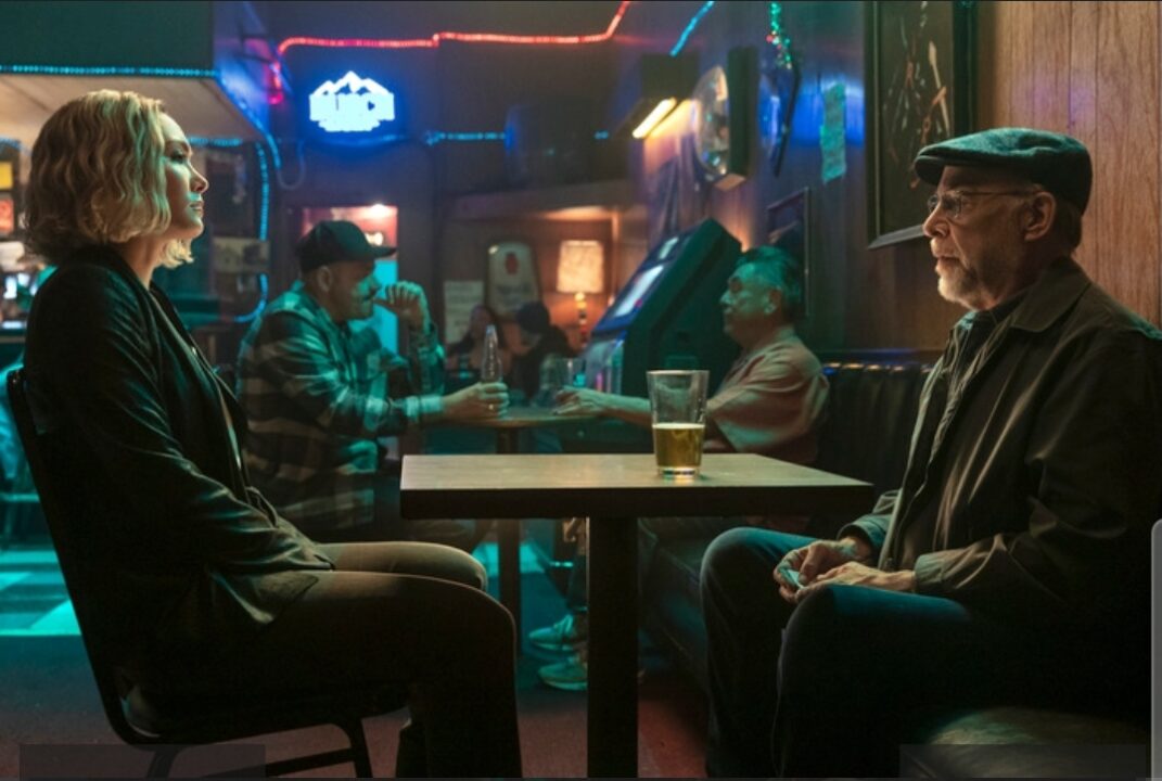 Raymond King (J.K. Simmons) in a diner booth, sitting across from Anaรฏs, in a dimly lit, blue-hued setting.