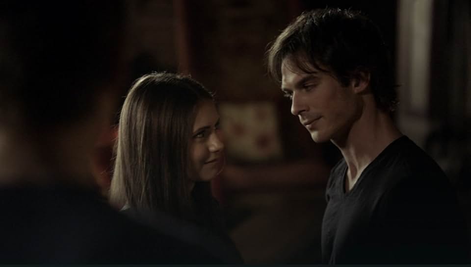Ian Somerhalder as Damon Salvatore looking at Elena Gilbert with a subtle smirk or knowing expression, in a dimly lit setting.