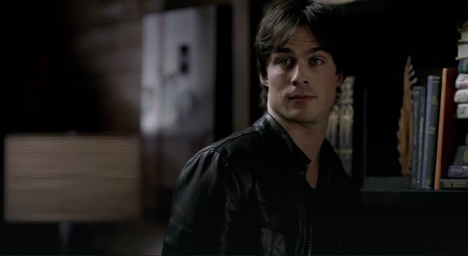 Ian Somerhalder as Damon Salvatore looking over a bookshelf in a dimly lit, possibly library-like room.