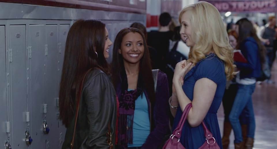 Elena Gilbert, Caroline Forbes, and Bonnie Bennett chatting by lockers in a high school hallway.