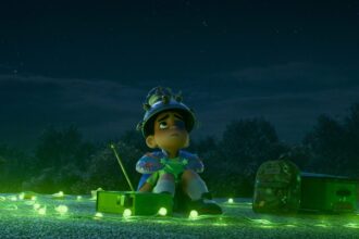 Elio, a young boy, sits on a rooftop at night surrounded by glowing green lights, looking up at the sky with a makeshift helmet, yearning for alien contact.