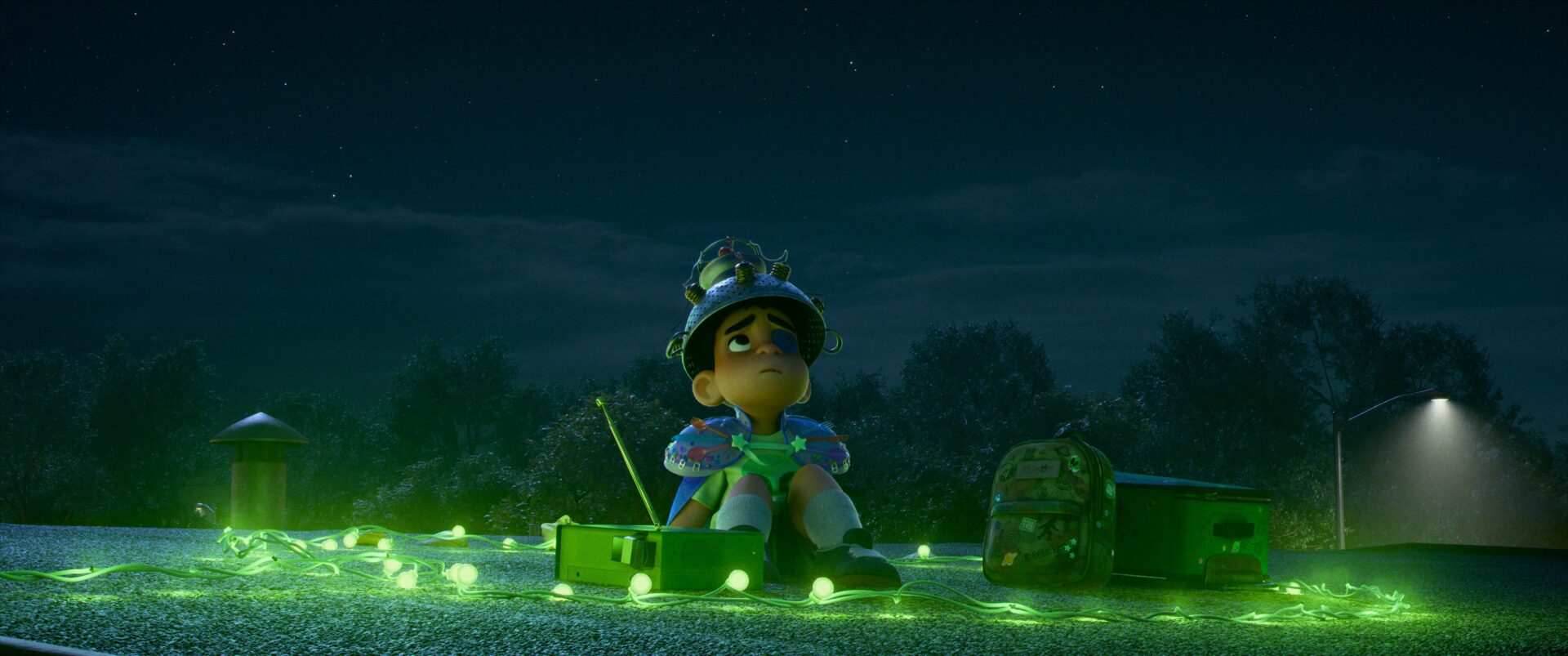 Elio, a young boy, sits on a rooftop at night surrounded by glowing green lights, looking up at the sky with a makeshift helmet, yearning for alien contact.